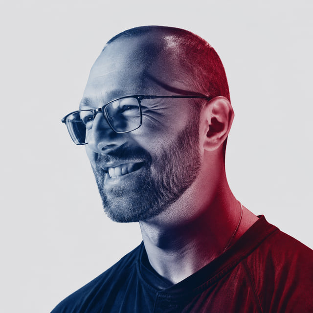 Jesse Jusek, smiling and looking off camera, in a duotone portrait lit deep blue on one side and red on the other